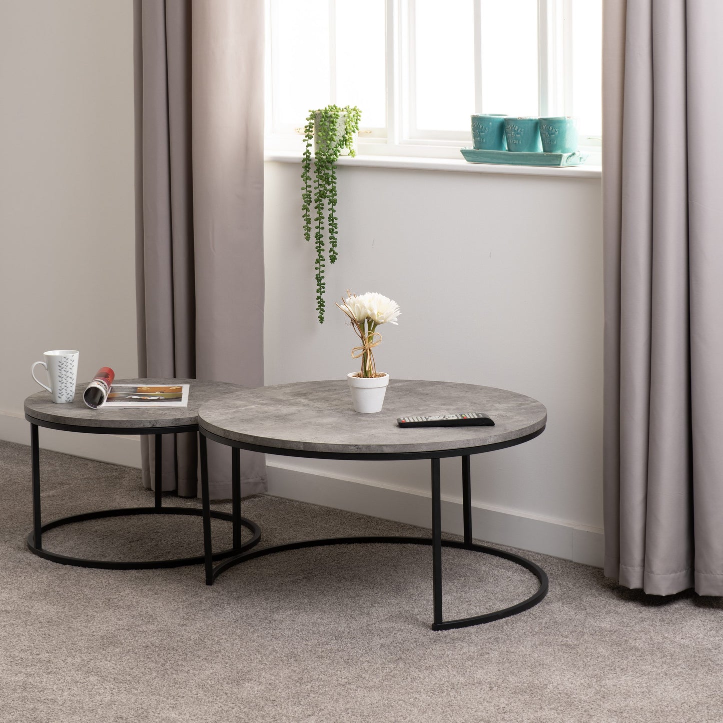Two round coffee tables with a gray concrete top and black metal frame in a room with curtains and a window.