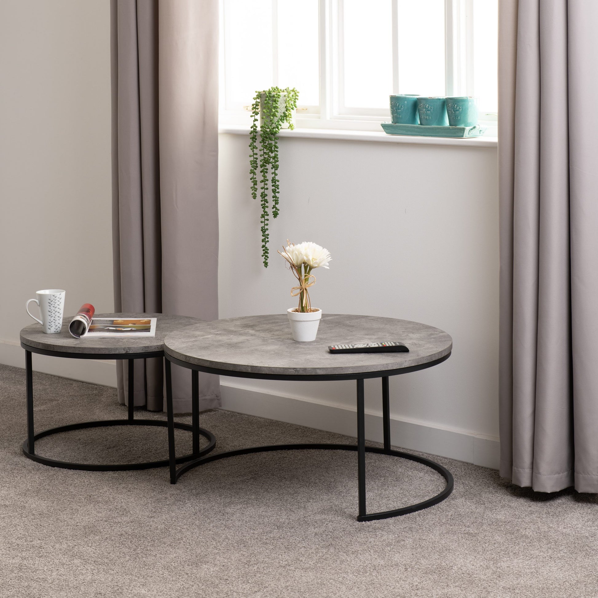 Two round coffee tables with a gray concrete top and black metal frame in a room with curtains and a window.