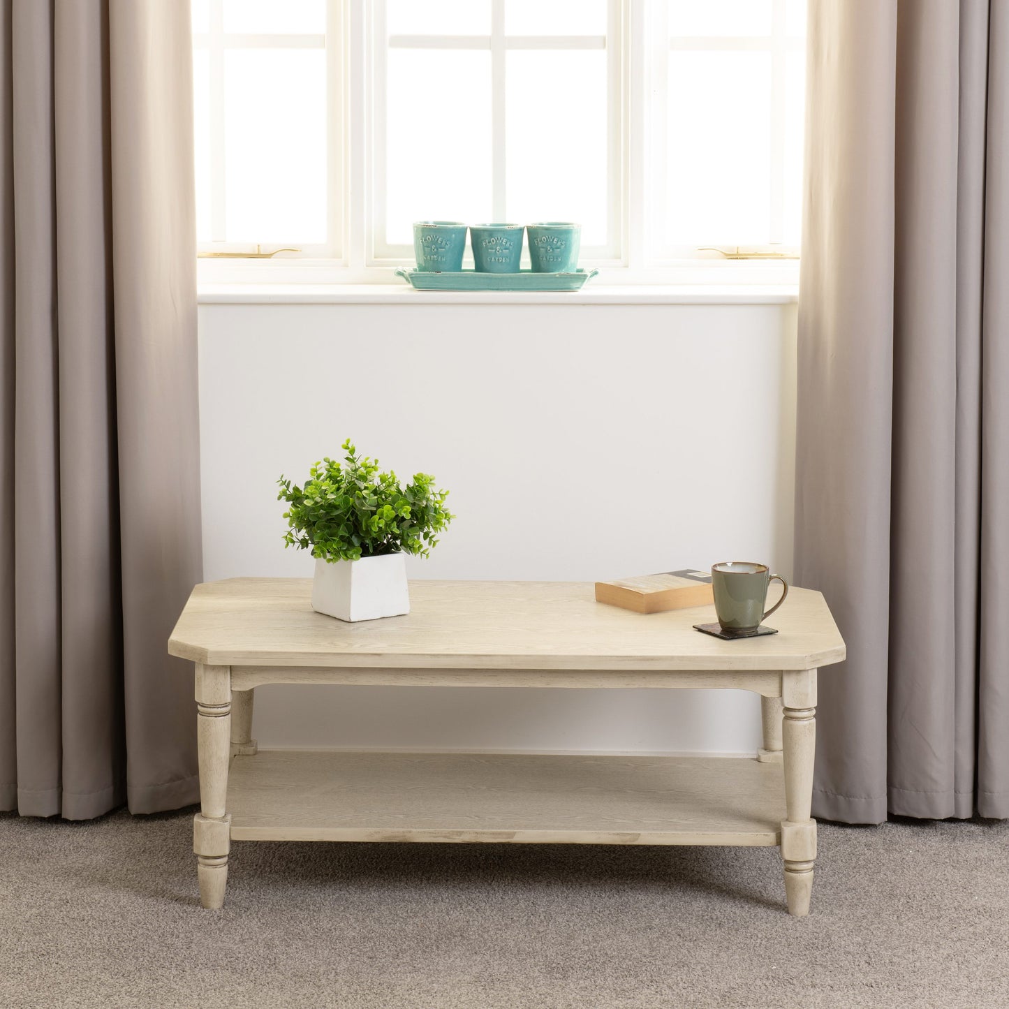 Light wooden coffee table with a plant and mug in a room with beige curtains and a window.