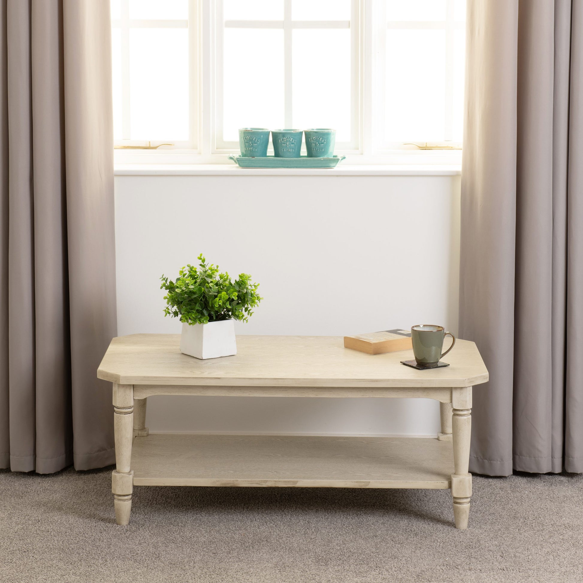 Light wooden coffee table with a plant and mug in a room with beige curtains and a window.