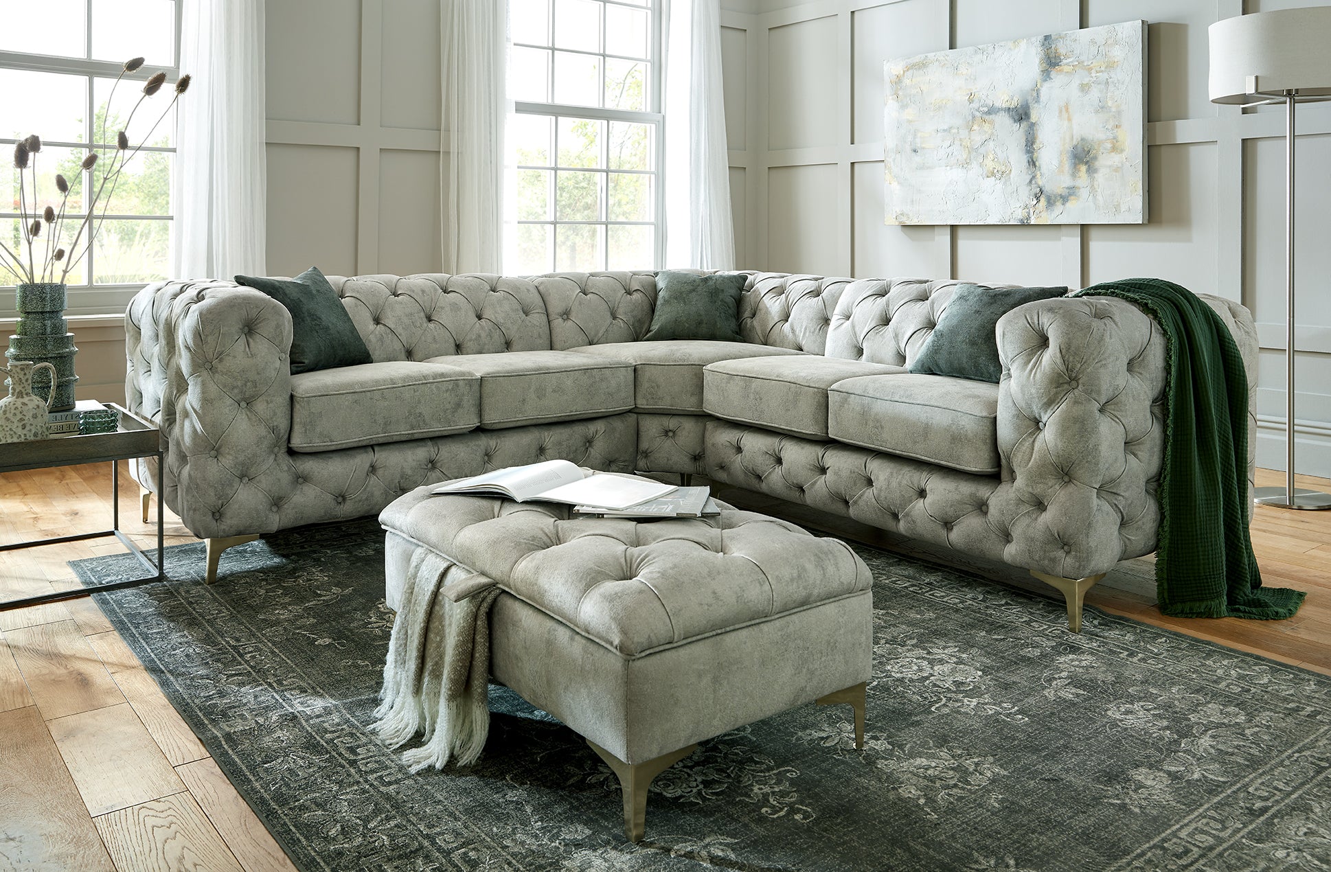 A large beige fabric corner sofa with tufted backrest and seat cushions, accompanied by a matching footstool, set on a patterned rug in a room with a painted wall and a window.