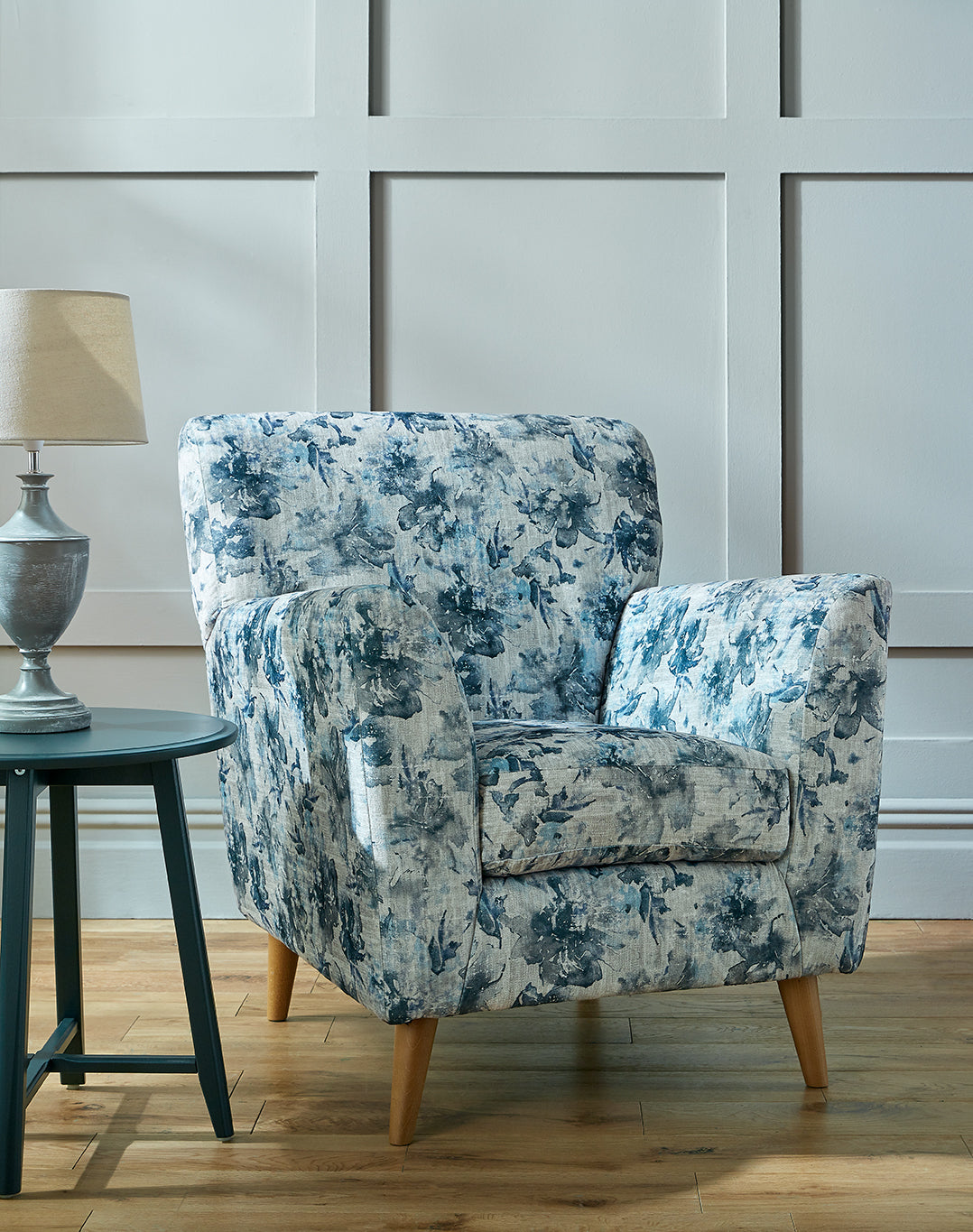 Blue floral armchair in a room with a wooden floor and a lamp on a table.