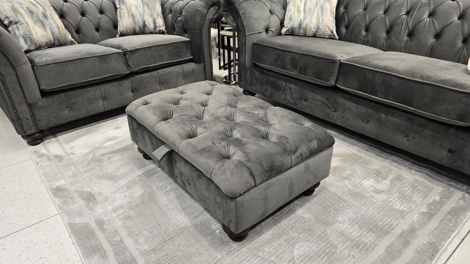 Gray tufted ottoman in front of a gray sofa set on a patterned rug.